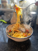 A close-up of a bowl of pasta with vegetables and meat, with a fork lifting a strand of spaghetti above the bowl. The background is a blurred kitchen setting.