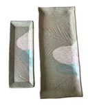 Two rectangular ceramic serving trays with a speckled sage green glaze and abstract painted motifs in pale pink, white, and light blue, shown on a plain white background.