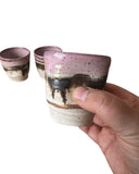 A photo of three ceramic cacao cups in beige, bronze, pink and gold. One cup is being held. 