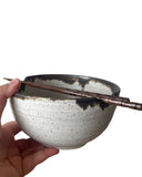 A handmade ceramic bowl with a speckled off-white glaze and a dark brown rim, held by a hand on the left with a wooden chopstick resting across the top, against a plain white background.