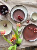A styled tabletop arrangement of handmade ceramic dishes on a beige linen cloth, with red grapes, plums, tulips, and a small cream cup.