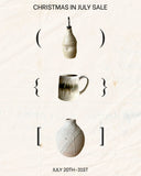 A promotional sale graphic on a textured off-white background with the text 'CHRISTMAS IN JULY SALE' at the top and 'JULY 20TH-31ST' at the bottom. Three ceramic home items are shown with bracket symbols around them: a small hanging bottle or vase at the top, a handled mug in the middle, and a rounded vase at the bottom.