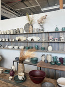 A collection of handmade ceramic tableware, including bowls, mugs, and vases, displayed on rustic wooden shelves and tables in a pottery studio.