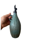 A hand holding a teal green ceramic oil pourer on a white background. The pourer is carved with a black metal weighted pourer. 