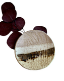 A handcrafted ceramic ring dish with a textured surface and a subtle striped pattern in beige, pink, and bronze colors, displayed with dark leaves in the background.