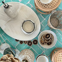 A styled flat-lay of ceramic home decor and serving vessels on a turquoise tiled surface, including a large leaf-shaped tray, speckled bowls, woven baskets, and dried citrus slices.