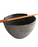 A beige and brown stoneware noodle bowl with a satin matte finish and bronze accent at the lip, accompanied by a pair of chopsticks resting on the bowl.
