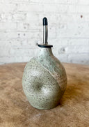 Speckled ceramic oil bottle with a metal nozzle on a wooden surface.