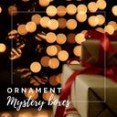 Promotional graphic for an 'Ornament Mystery Box' featuring wrapped gift boxes with red ribbons against a background of warm golden bokeh lights, enclosed in a thin white square frame with white text overlay.