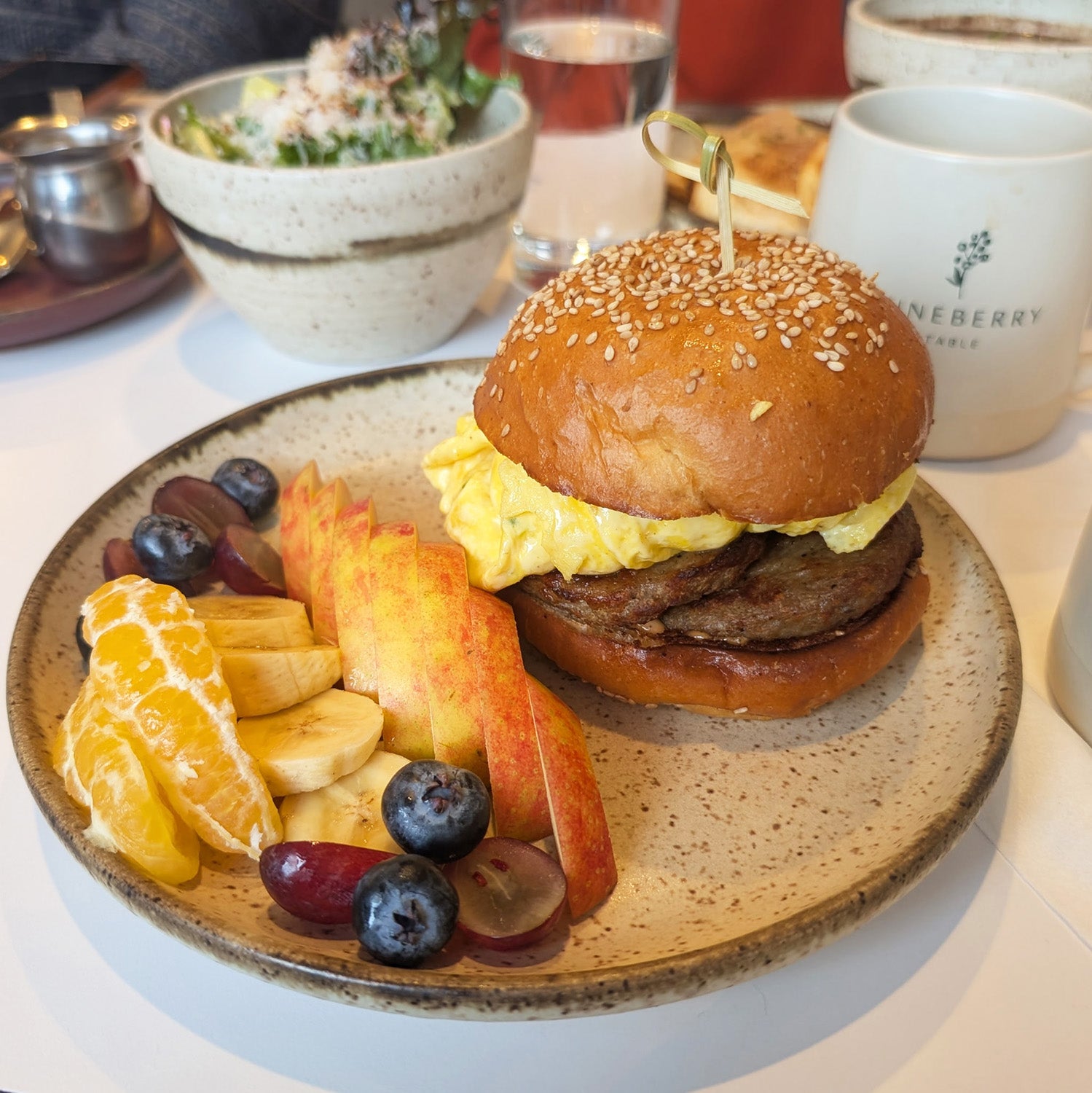 A breakfast sandwich and fruit on a handmade plate with food in the background. 