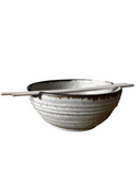 A set of handcrafted ceramic bowls in various colors, with a rustic texture, stacked together with a pair of chopsticks resting on top.