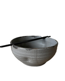A set of handcrafted ceramic bowls in various colors, with a rustic texture, stacked together with a pair of chopsticks resting on top.