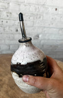 Hand holding a ceramic bottle with a black and white speckled design against a white brick wall.