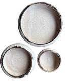 Three round ceramic bowls in different sizes arranged on a plain white background, shown from above. The bowls have a speckled off-white interior glaze with a contrasting dark brown to black rim.