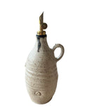 A beige and bronze handmade olive oil bottle with a gold weighted pouring spout on a white background. 