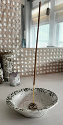 Incense stick in a speckled ceramic bowl against a textured wall.