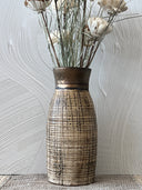 A rustic ceramic vase with a textured, grid-like pattern in beige and brown, featuring a metallic bronze-toned neck, filled with dried white flowers and placed on a textured gray surface.