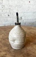 A handmade ceramic oil dispenser bottle with a speckled off-white glaze and a black pour spout, set on a rustic wooden surface against a white brick wall background.