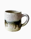 A handcrafted ceramic mug with a unique glaze resembling the Aurora Borealis, with beige, bronze, white, and pink hues.