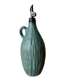 A teal green ceramic oil pourer on a white background. The pourer is carved with a black metal weighted pourer. 