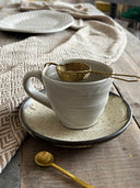 A ceramic up with a tea strainer on a plate with a table cloth on a rustic wooden table. 